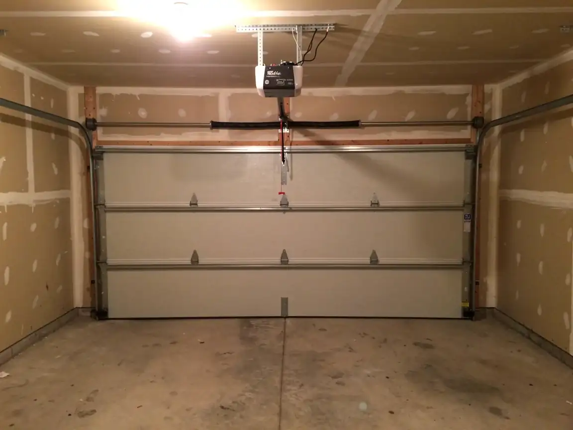 A white garage door is closed inside a garage with unfinished drywall.